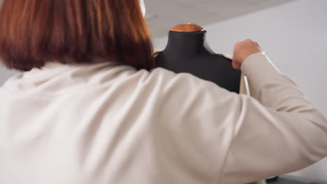 garment maker puts beige sleeveless blazer onto black dress form, guiding blazer into place and fastening single front button, adjusting shoulders and seams for fit and precision in fashion workshop