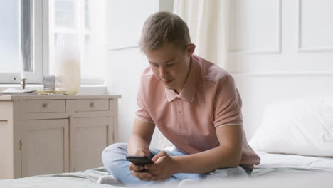 boy with down syndrome using smartphone sitting on the bed in the bedroom at home