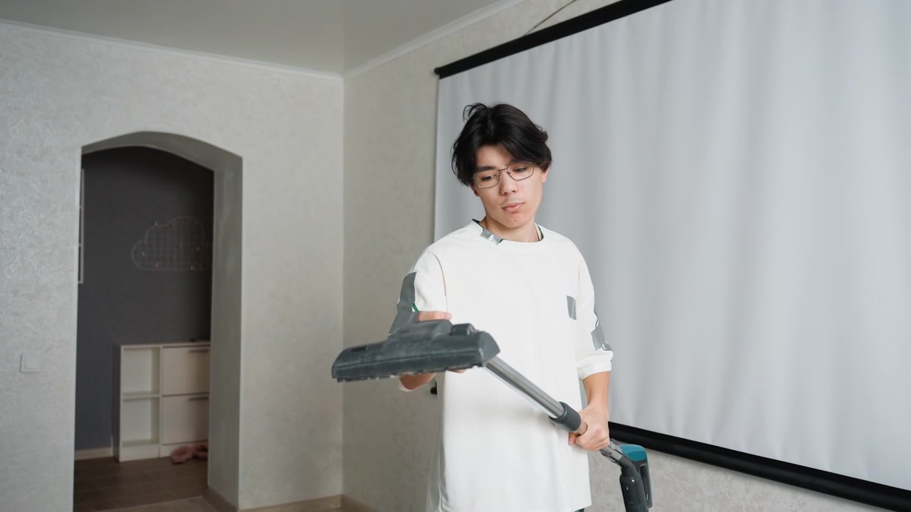 リビングルームを掃除機で掃除するアジア系の若い男性、カジュアルな白いシャツ、伸縮式のスティック掃除機、プロジェクタースクリーンを背景に、ニュートラルな壁とアーチ型の通路、規則的でゆっくりとした動き、家事の掃除ルーティン