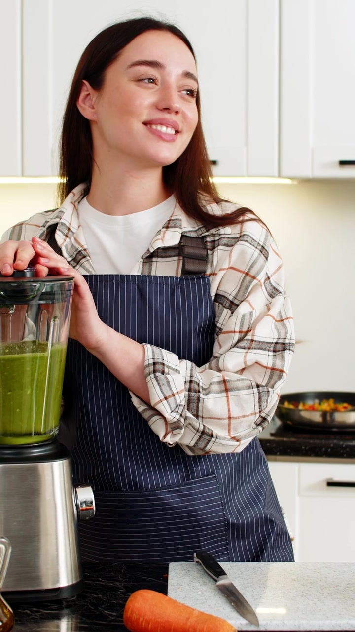 Giovane donna avvia il frullatore per un frullato verde con spinaci, banana, mela e carota in cucina.