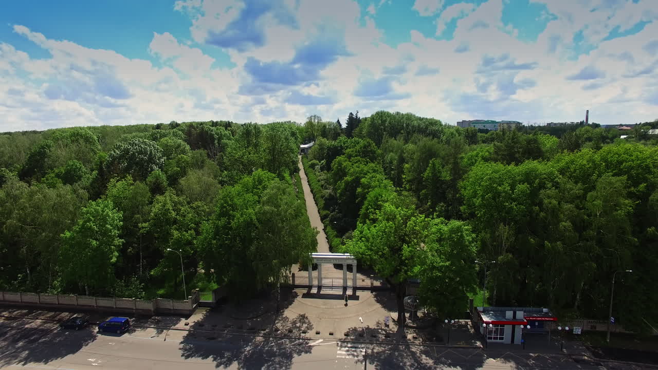Entrance of the green city park with long road in the middle. Drone footage rising over the crossroads on sunny day.