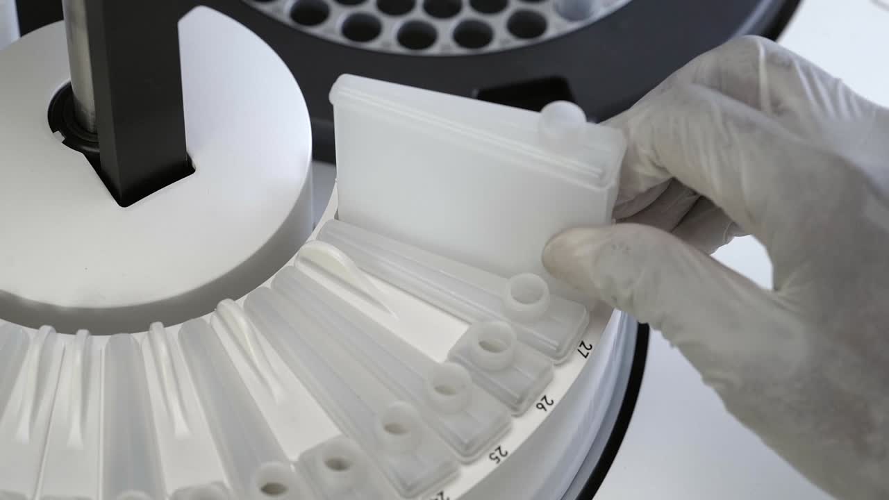 장갑을 입은 실험실 기술자가 화학 분석기에서 반응기 병을 제거합니다.