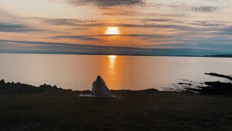 Beautiful-young-woman-sitting-on-blanket-on-the-top-of-mountain-cliff-in-front-of-the-Atlantic-ocean-at-sunset