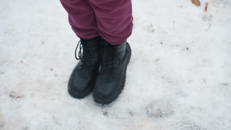 vista de ángulo inferior de un individuo con botas negras saltando juguetonamente en el suelo cubierto de nieve con follaje seco esparcido, centrarse en el calzado, el movimiento y el entorno de invierno