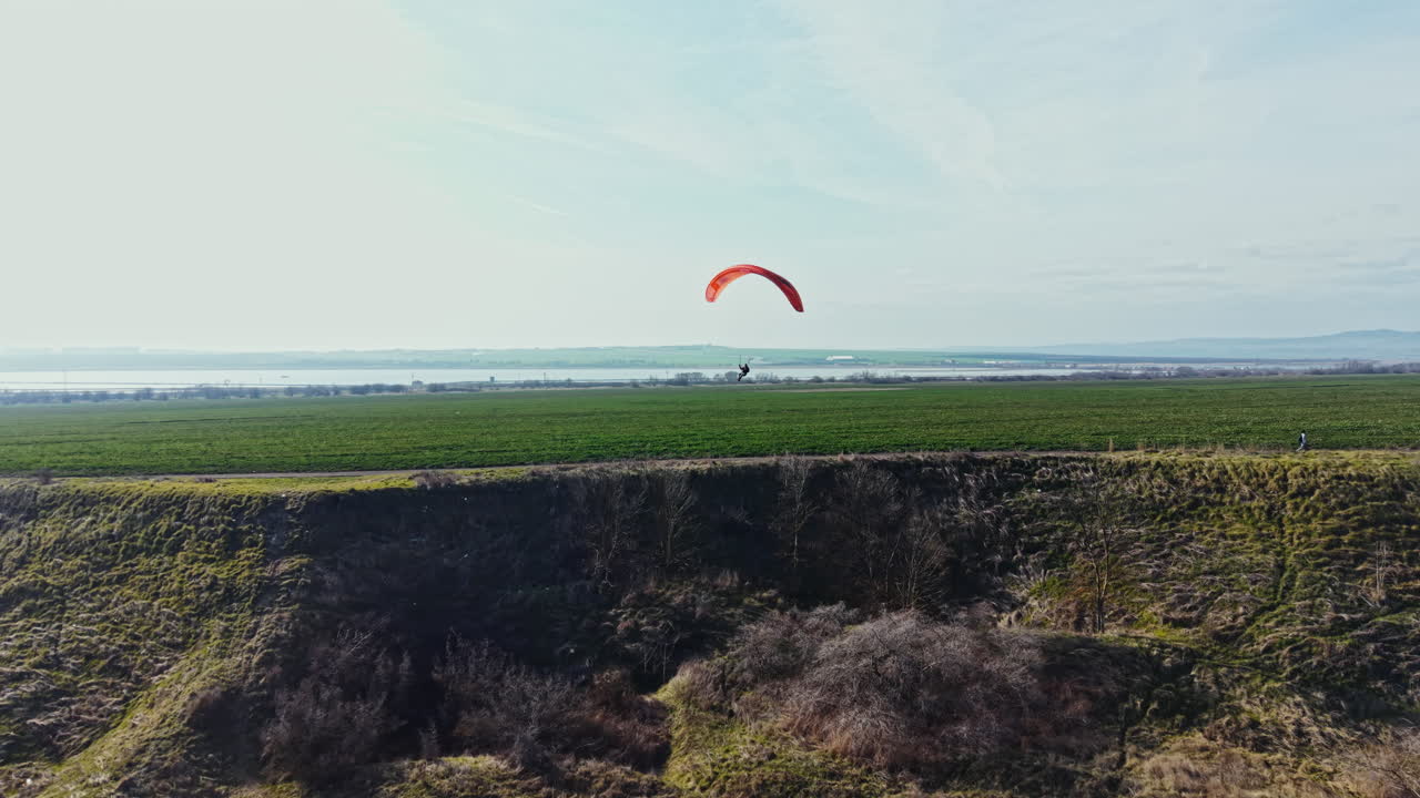 Parapente voa sobre campos verdes perto de um corpo d'água em um dia claro com céu azul e poucas nuvens.