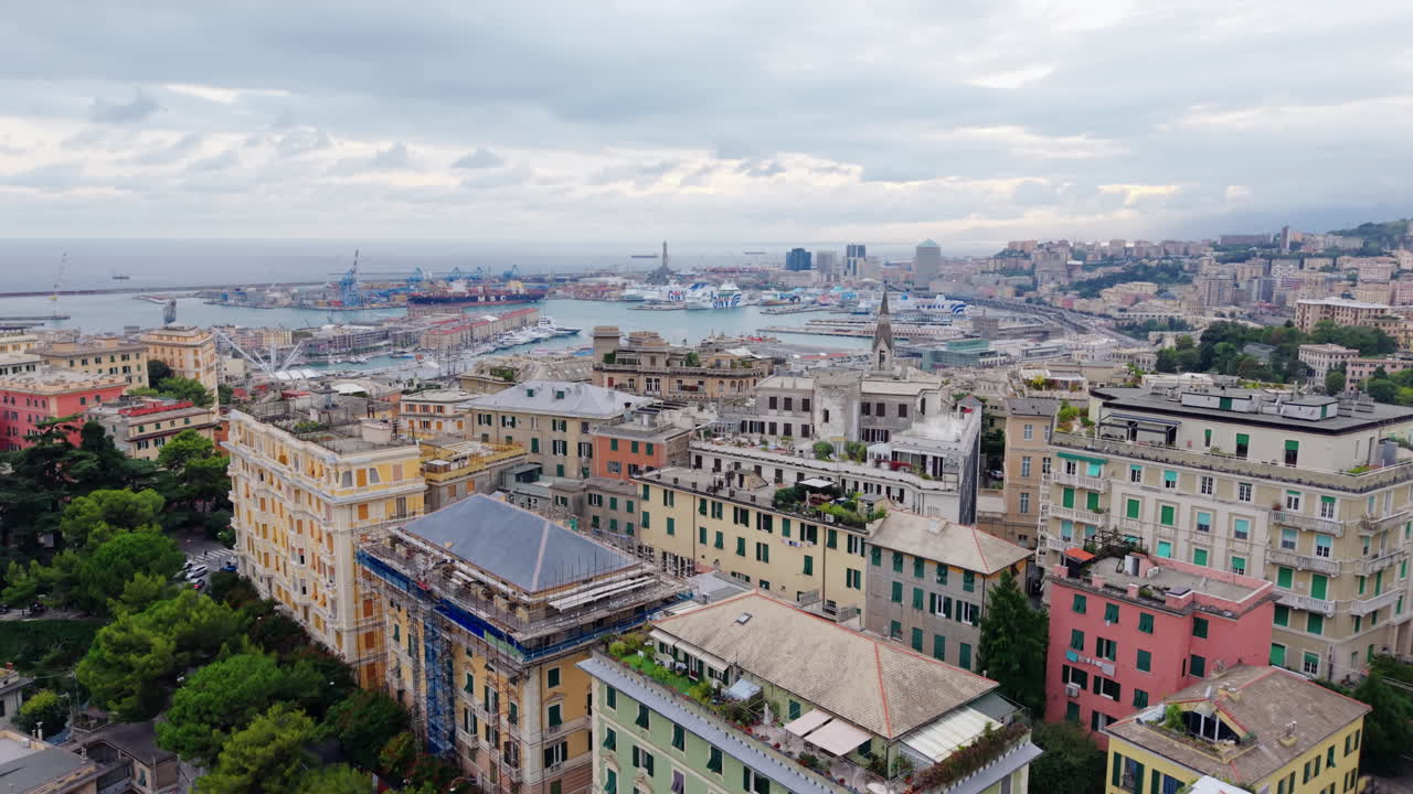 A drone ascends over colorful residential buildings and trees, revealing the historic center of Genoa and its port area with ships and cranes under a cloudy sky