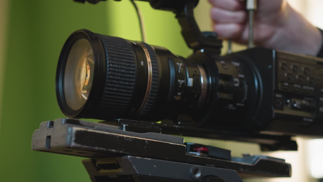 Close-up of camera lens mounted on tripod, with hand adjusting position. Background features a green wall and blurred elements, creating a dynamic, cinematic scene