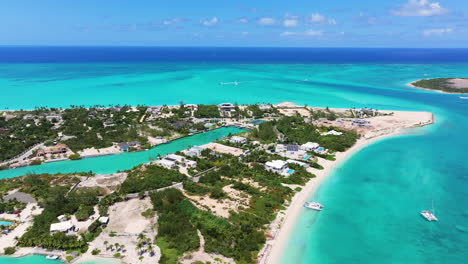 Upward slide shot of coastal land nestled between canal and vast ocean, revevealing villas and greenery lining the shores. The clear blue water of the canal merges seamlessly with the turquoise ocean