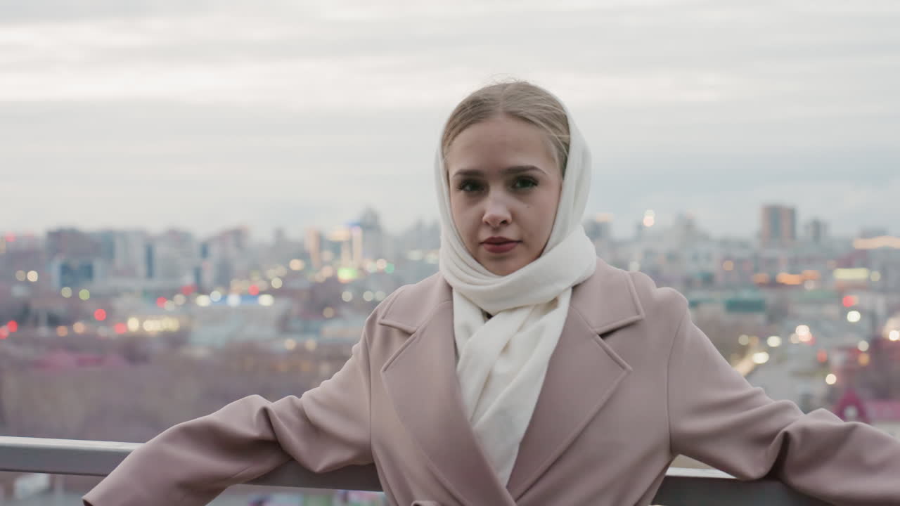 Donna caucasica con le braccia aperte contro lo skyline della città al tramonto, cappotto e sciarpa svolazzanti, modella sicura in posa per un servizio editoriale serale, luci panoramiche e orizzonte calmo amplificano il senso di forza.