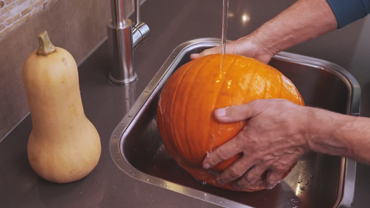 Man rinses bright orange pumpkin under running water in kitchen sink, preparing for seasonal cooking