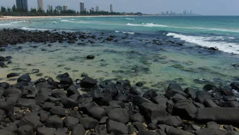 Burleigh-Heads,-Black-Rocky-Shore,-Distant-City-Skyline,-Surf-AERIAL