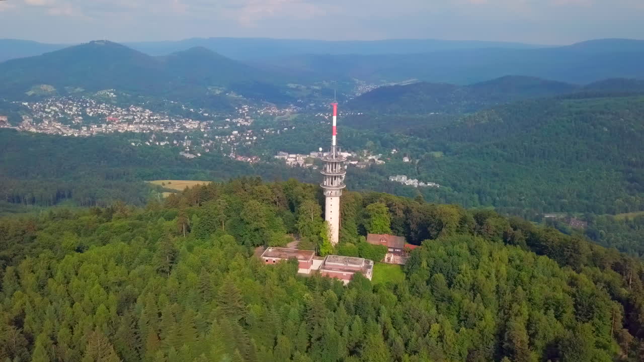 Surrounding aerial view of the 'Fremersberg' transmission tower on a green forested mountain with the spa town of Baden-Baden in the background on a sunny summer day.