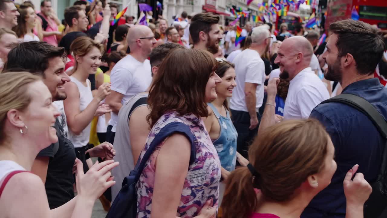 Festival di strada vivace con folla eterogenea, bandiere arcobaleno e autobus a due piani.
