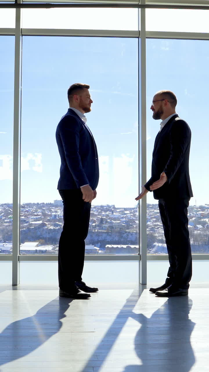 Businessman greeting in office. Top managers in business suits, shaking hands with each other, against the panoramic window view with a city background. Vertical video