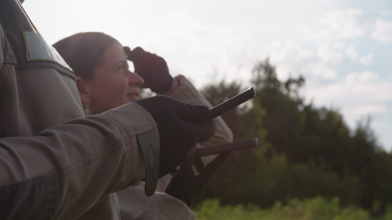 close up of person holding remote controller while female partner gazes skywards, gloved hands gripping transmitter, sun flare and device detail framed against forest under cloudy sky