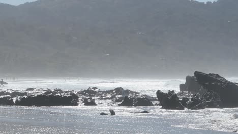 Surfer-coming-out-of-the-beach-into-the-sand-as-small-waves-crash-on-the-rocks-during-daylight