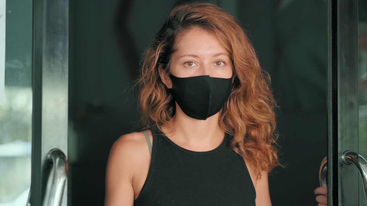 Kvinna med ansiktsmask bakom glasdörr med karantänskylt.