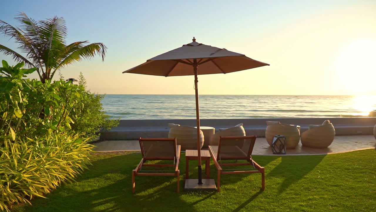 Empty chairs under outdoor umbrella overlooking a peaceful ocean scene at sunset. Calm ocean reflecting light of the setting sun. Luxury island resort furnishings. Relaxing setting no people