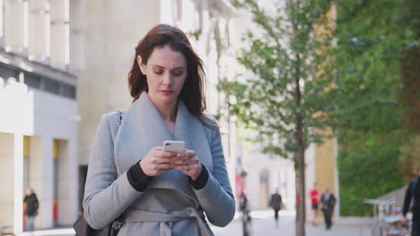 young white woman walking in a sunny london street stops and uses her smartphone, close up