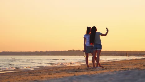 young women posing to camera on the phone at the seaside at sunset. two girls taking selfie in mobile phone in the evening sea.