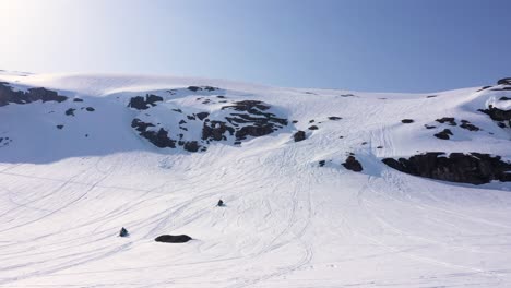 Aerial view over snowy mountain landscape. Snowmobiles driving up mountain side. Sunny weather. MOVING RIGHT TO LEFT.