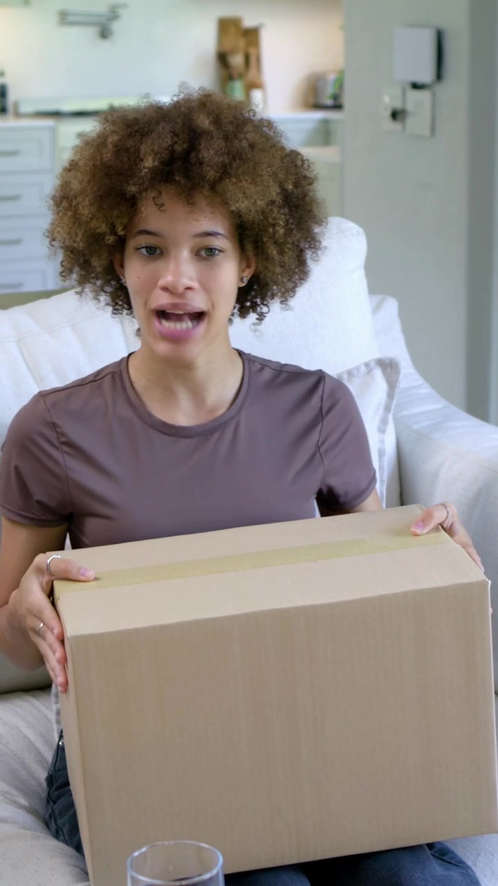 Vídeo vertical: Reagindo a uma voz fora da tela, amigos com blusas bordô e taupe manuseando uma caixa no sofá.