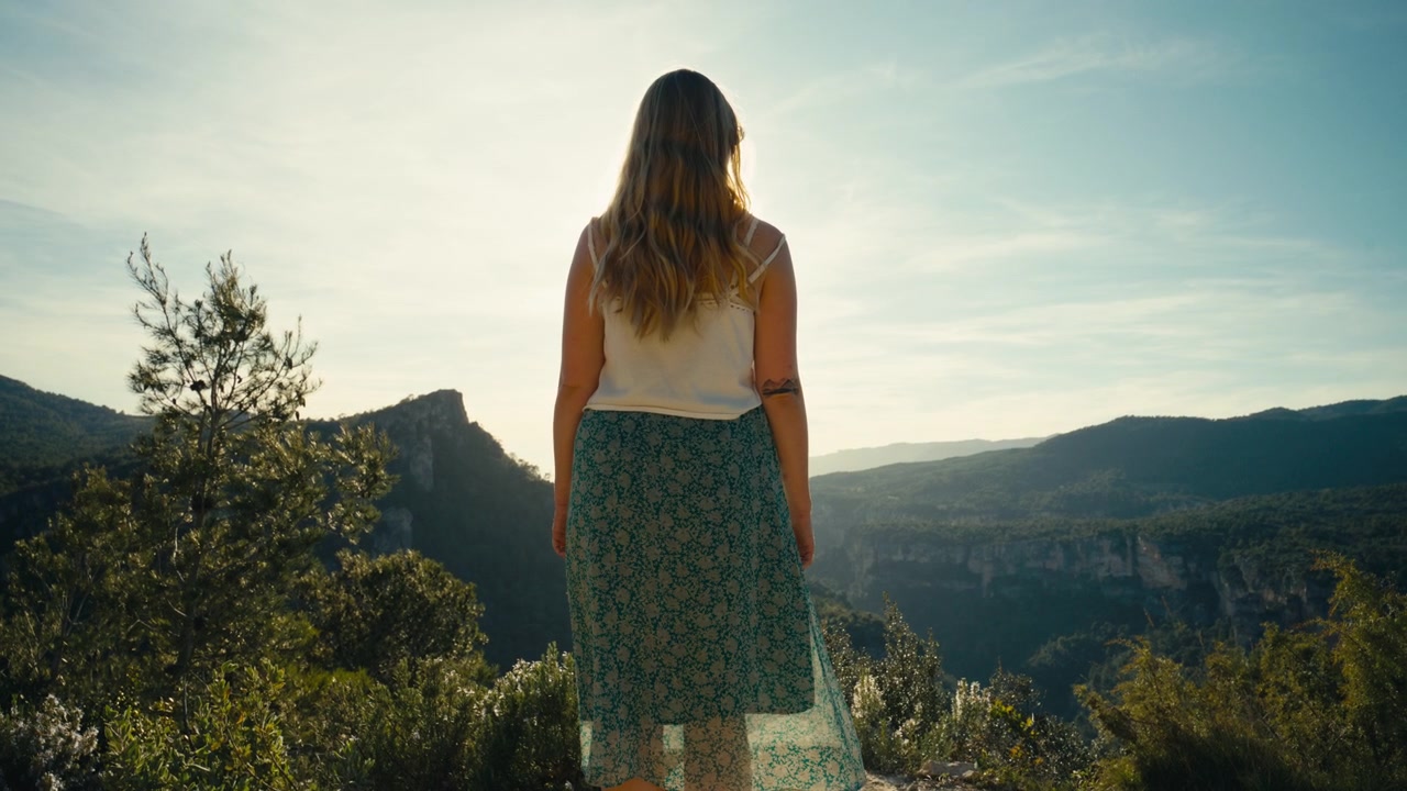 A woman stands still above a canyon in Catalonia, Spain, facing the fading light as cliffs, trees, and distant ridges sink into a soft backlit haze.