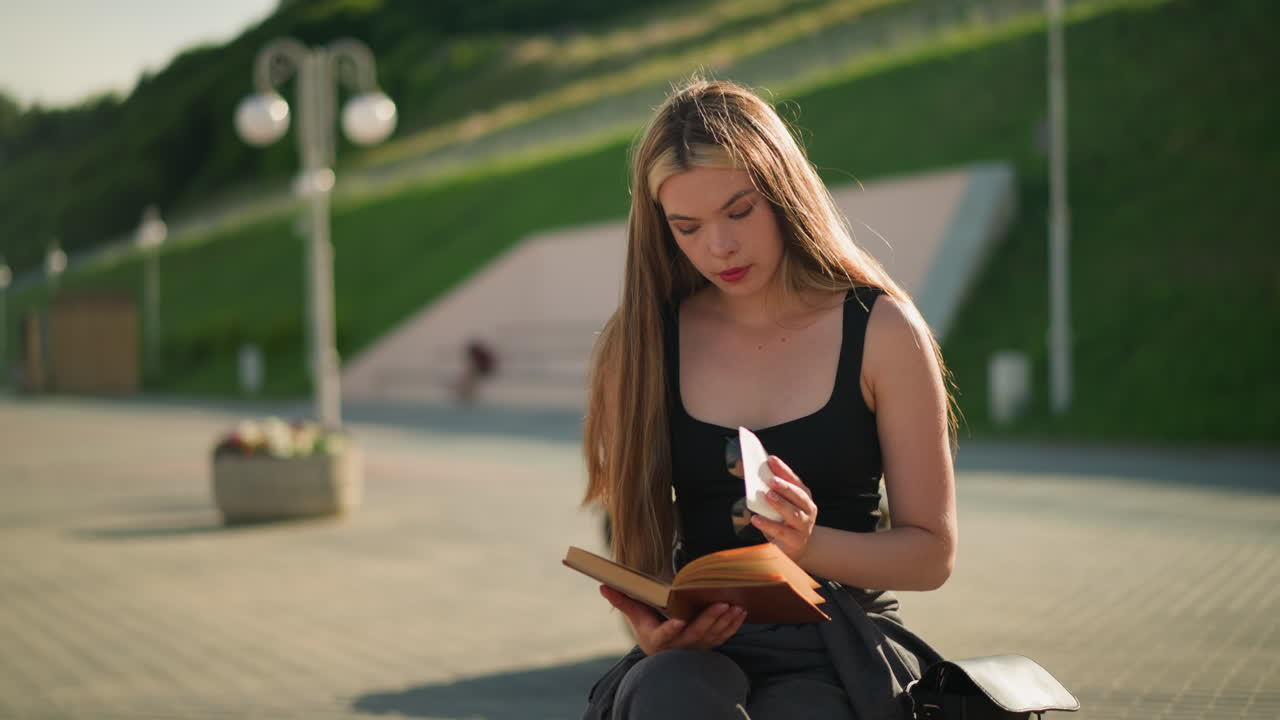 Woman seated outdoors on a bench with legs crossed, flipping through pages of her book and contemplatively observing a piece of paper, the background features lamp poles