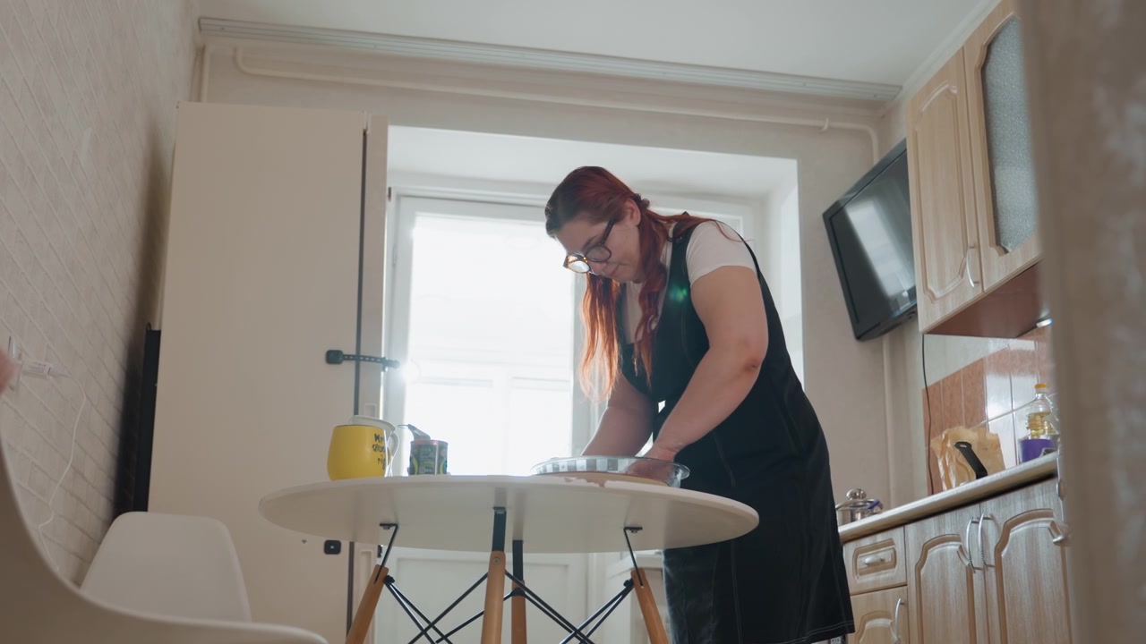 Young woman leans over round table in compact home kitchen, scrubbing plate with soap and running water, focused posture and bright window light convey routine household chore, with woman, kitchen