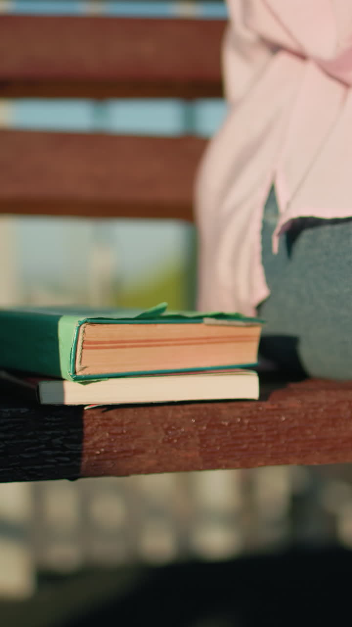 vista parcial de un individuo en vaqueros y camiseta rosa sentado en un banco con libros a su lado, escena al aire libre con luz solar cálida, fondo borroso con barandilla de hierro y vegetación