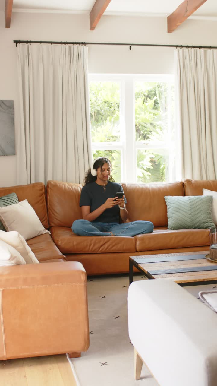 Vertical video: African American woman checking smartphone and hearing audio via headphones on sofa