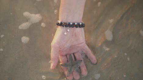 Mano-Femenina-Con-Una-Pulsera-Marrón-Y-Blanca-Sosteniendo-Una-Estrella-De-Mar-En-Agua-De-Burbujas-En-Movimiento-En-La-Orilla