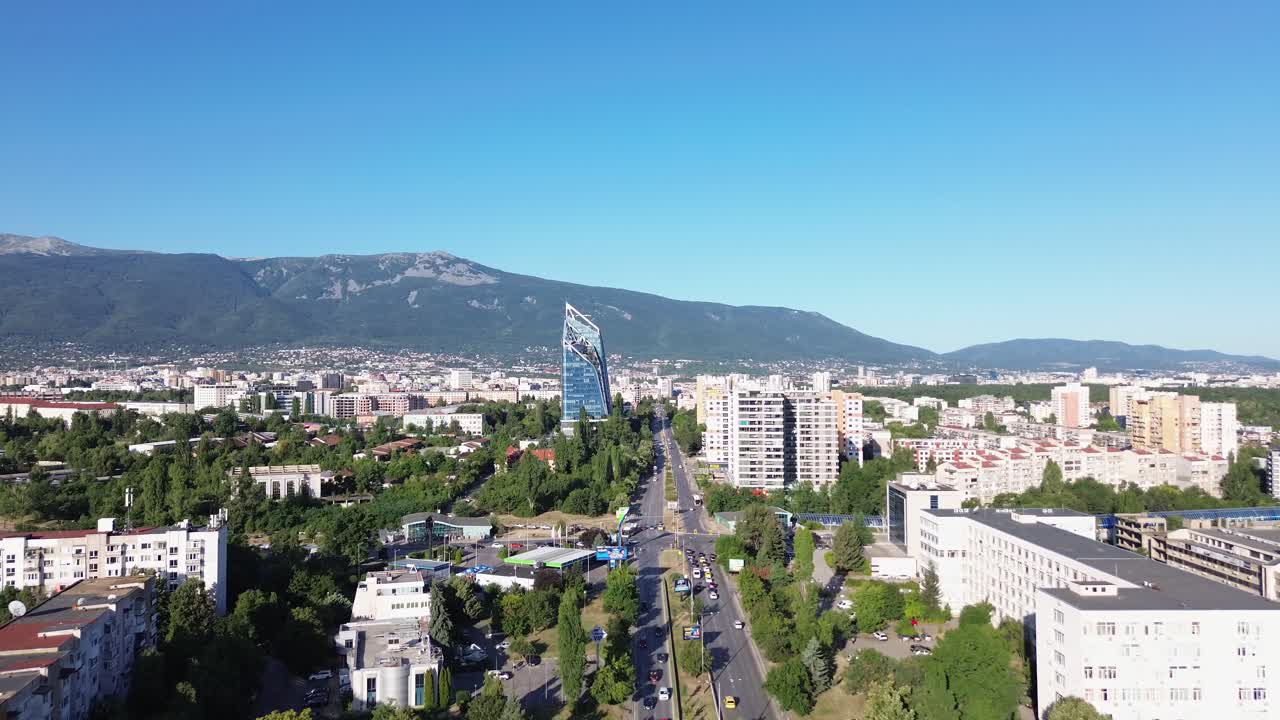Establishing aerial shot of a street in Sofia, Bulgaria, on a sunny, summer day with a clear sky and Vtosha Mountain behind. Some traffic on the street, big office building standing out.