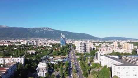 establishing aerial shot of a street in sofia, bulgaria, on a sunny, summer day with a clear sky and vtosha mountain behind. some traffic on the street, big office building standing out.
