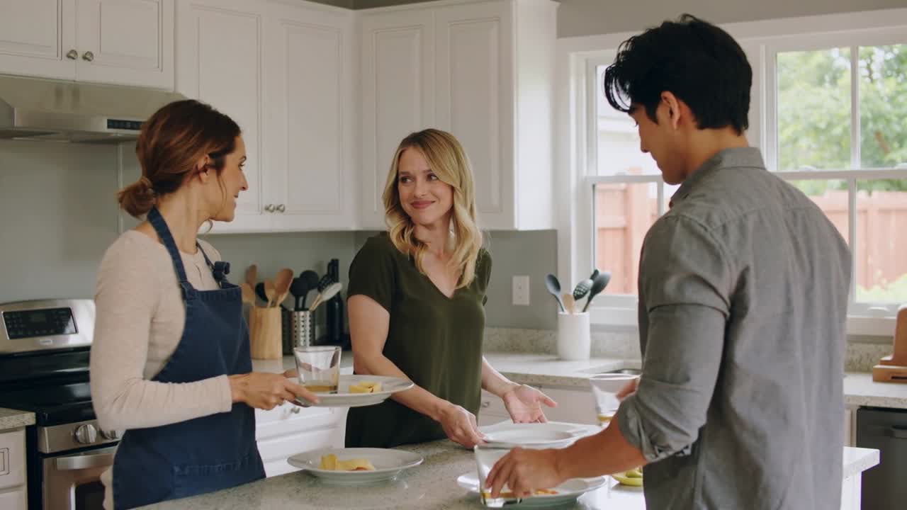 Left woman bringing tray toward island center woman catching plates man plating food preparing meal
