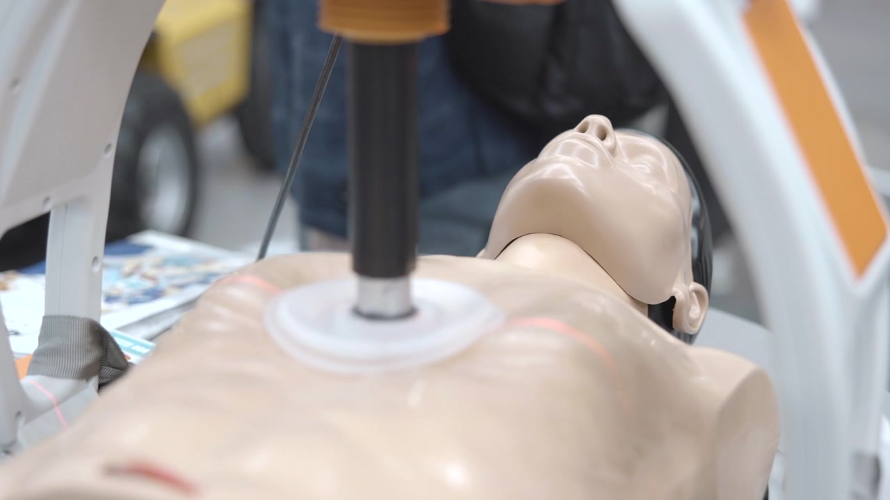 Voksen træningsdukke ligger på et medicinsk bord, mens et kompressionsapparat presser torsoen i et klinisk simuleringsrum, med glat ansigtsmaske, støttestativ, sløret udstyr, rene overflader og lyst