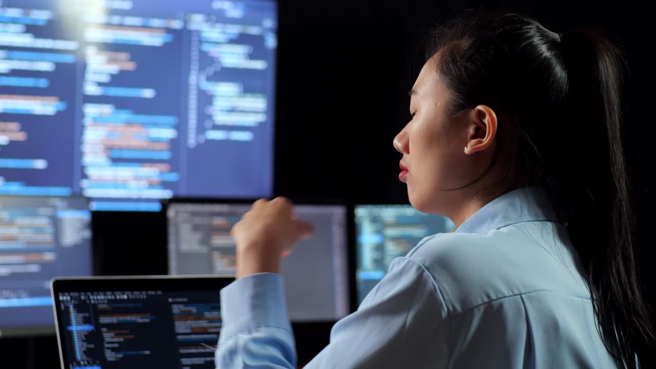 vista de cerca de atrás de una programadora asiática pensando en algo mientras escribe código por una computadora portátil usando múltiples monitores que muestran la base de datos en terminales ventanas de escritorio en la oficina