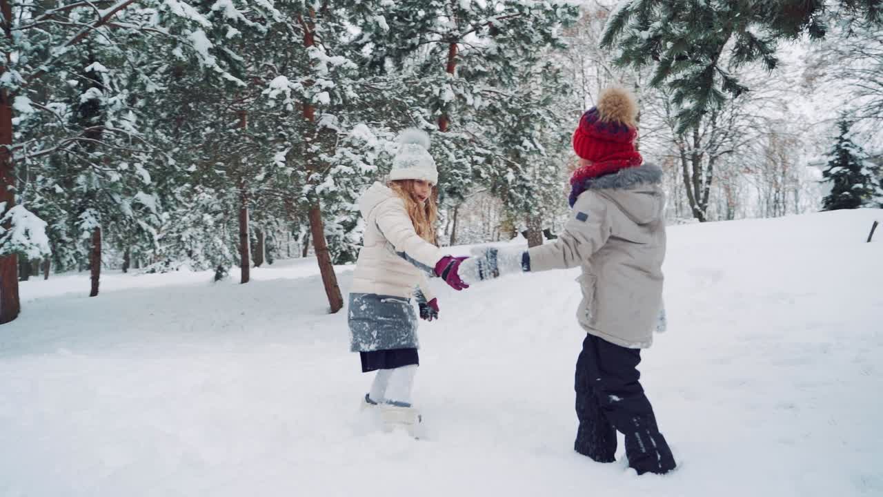 Children are holding each other's hands in the yard and running up in a snowy hill together on the background of trees in the winter. Slow motion