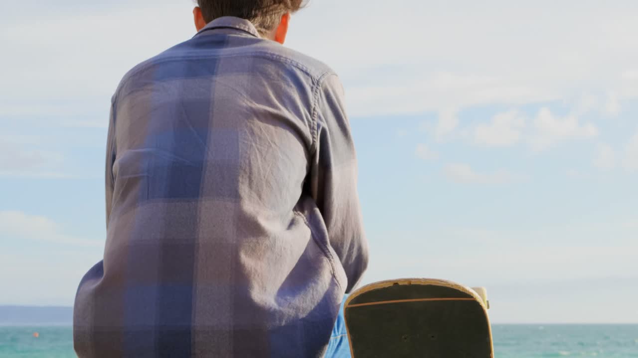 해변에서 바다를 바라보는 스케이트보드와 함께 젊은 백인 남자의 뒷면 4k