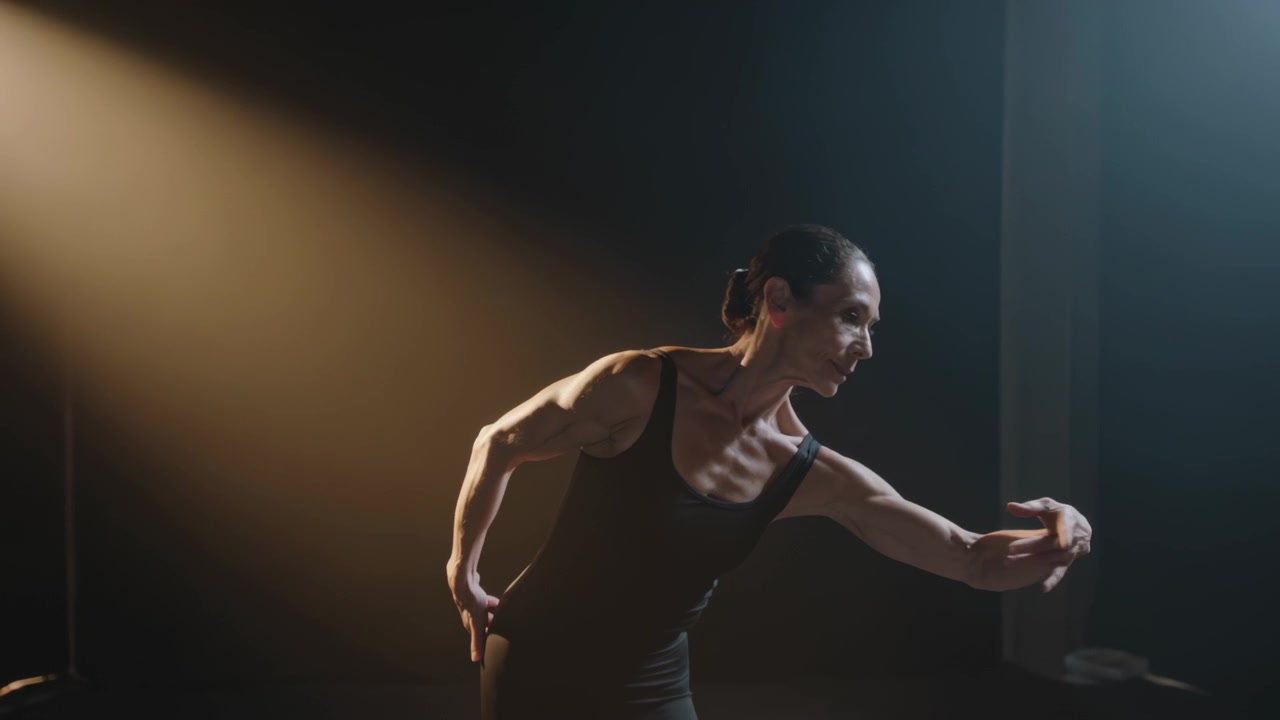 Reaching on spotlight cue, dancer in black leotard performing solo, extending right arm on stage
