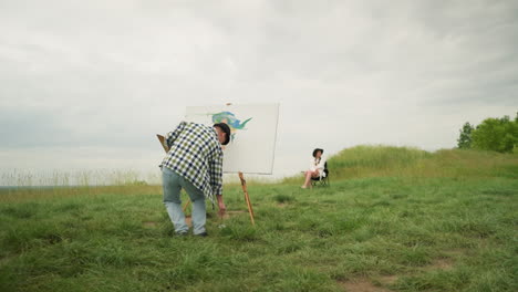 back view of an artist, wearing a hat and checkered shirt, focused on painting on a board in a grassy field under a cloudy sky. a woman, dressed in a white dress and hat, sits in front of him