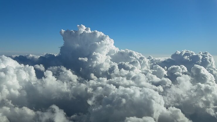 An immersive pilot’s eye perspecvtive taken from the cockpit of a jet airplane flying above a cottony storm clouds under a deep blue sky, with a threatening cumulonimbus storm cloud ahead