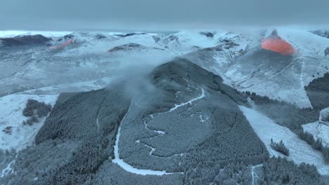 Sonnenaufgang-Mit-Orangefarbenem-Relief-Des-Bergschattens-Auf-Schneebedecktem-Gipfel-Mit-Frostigem-Kiefernwald,-Niedriger-Wolkendecke-Und-Entfernten-Schneebedeckten-Bergen