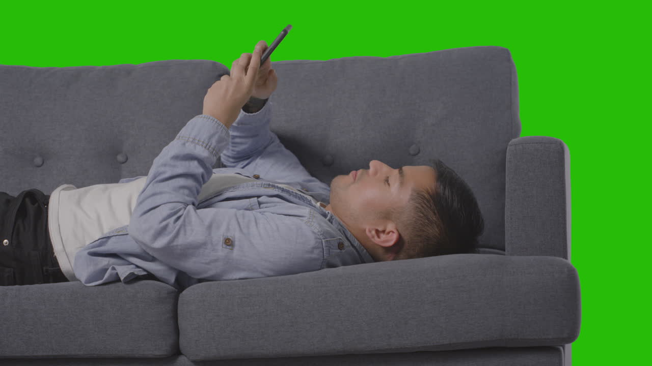 Studio Shot Of Man Lying On Sofa Scrolling Through Mobile Phone Against Green Screen