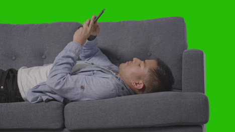 Studio Shot Of Man Lying On Sofa Scrolling Through Mobile Phone Against Green Screen
