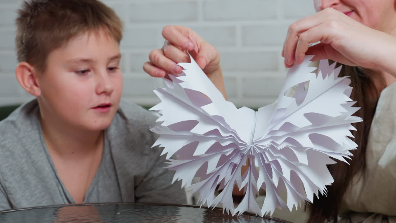 partial view of mother proudly holding handcrafted paper ornament while young son looks at artwork with wide-eyed amazement sitting together at table indoors with white brick wall in background