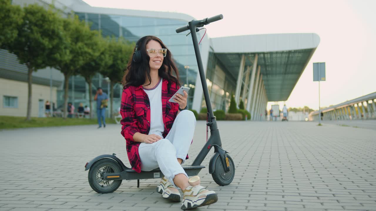 Front view of beautiful smiling satisfied carefree young brunette in glasses and trendy clothes which enjoying songs in earphones from phone playlist while sitting on e-scooter near urban modern building
