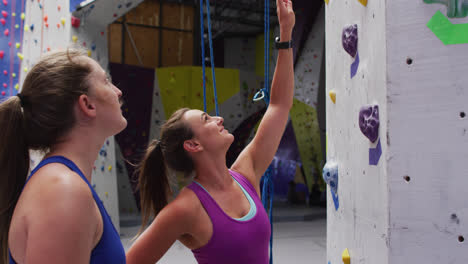 Dos-Mujeres-Caucásicas-Felices-Hablando-Y-Preparándose-Para-Una-Escalada-En-El-Muro-De-Escalada-Interior