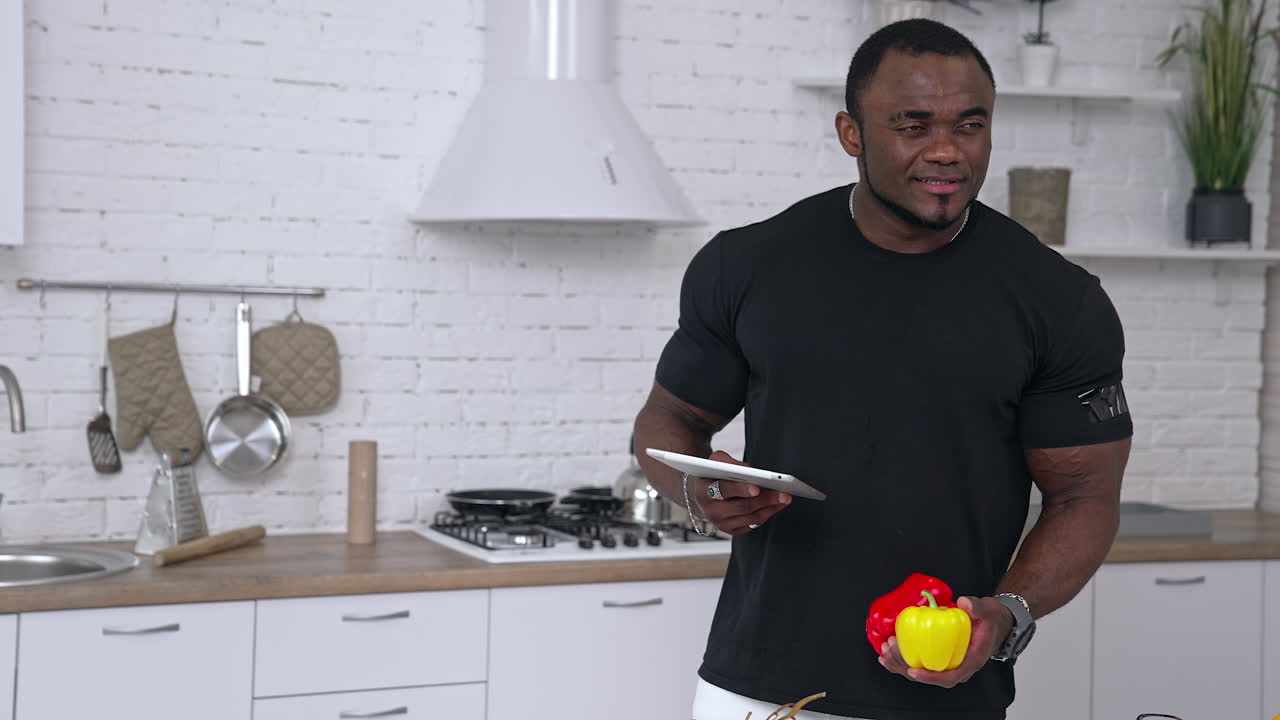 Afro-american sportsman in the kitchen. Portrait of a thoughtful black skinned man with muscular body holding tablet and fresh organic vegetables.