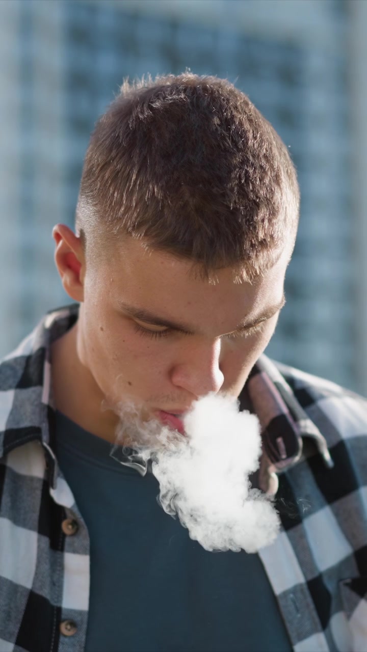 Close up of young man wearing casual clothing holding and using electronic vaping device outdoors. subject is seen exhaling vapor while maintaining minimal movement throughout scene